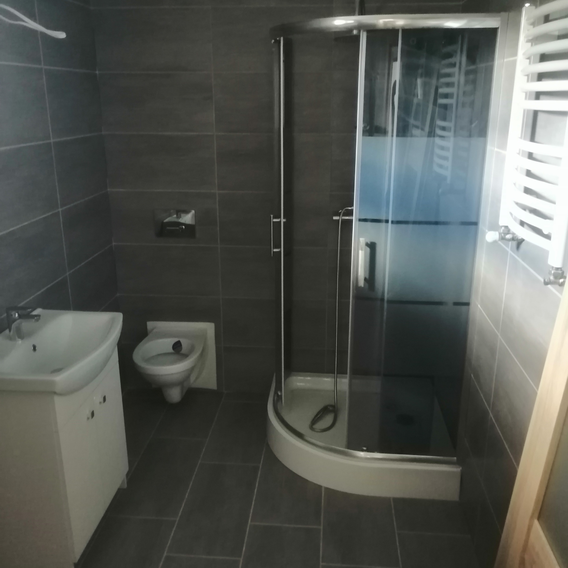 Szara łazienka z kabiną prysznicową z zaokrąglonymi drzwiami, umywalką z szafką, bidetem i grzejnikiem drabinkowym.