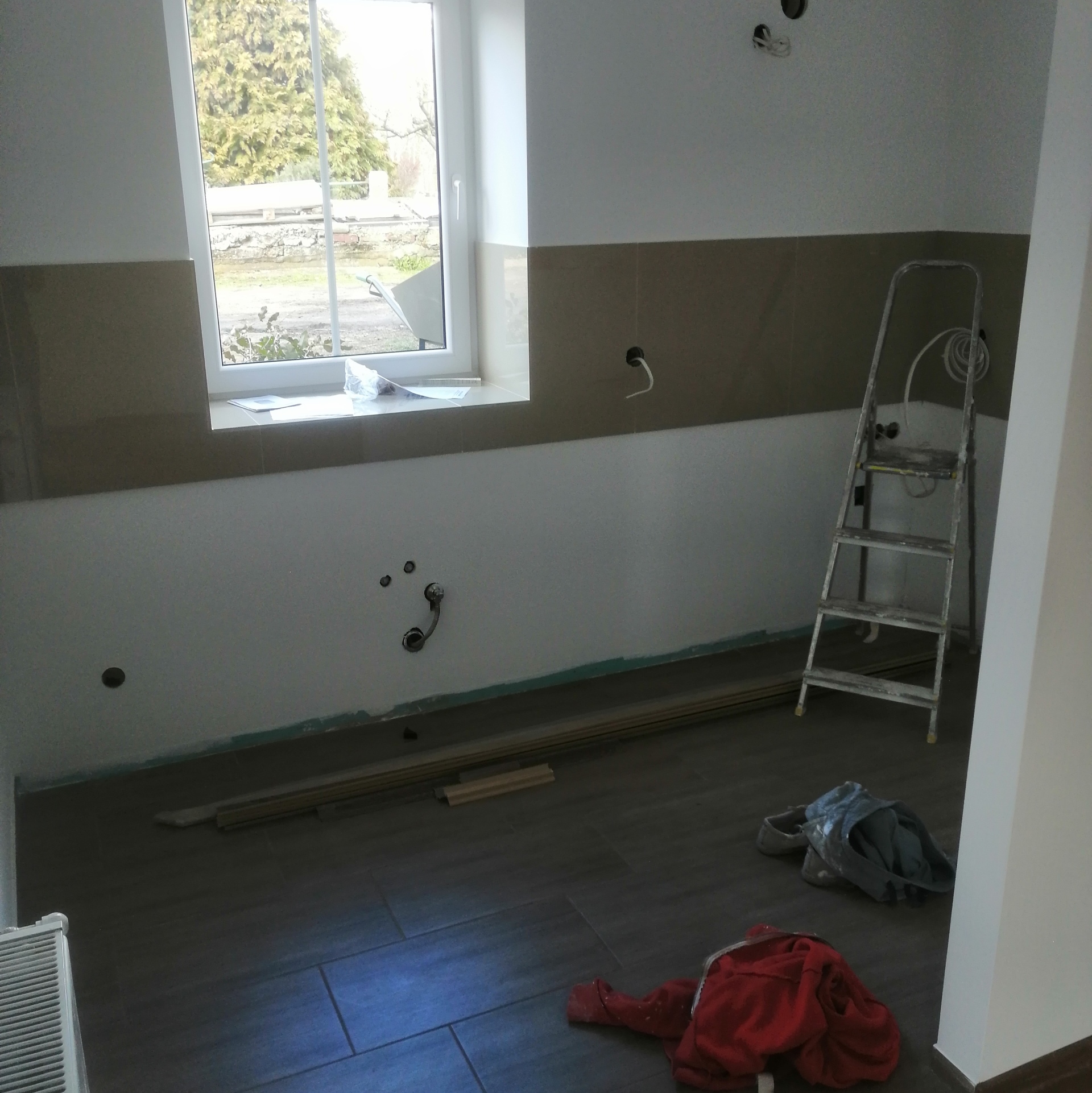 Wnętrze w trakcie prac wykończeniowych: ułożone płytki na podłodze i ścianie, widoczne przygotowane panele, drabina, instalacje elektryczne oraz hydrauliczne.