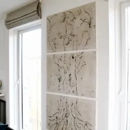 Nowoczesny salon z minimalistycznym wystrojem, na ścianie betonowy tryptyk przedstawiający stylizowany rysunek drzewa, obok okno z szarą roletą rzymską.