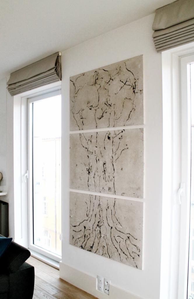 Nowoczesny salon z minimalistycznym wystrojem, na ścianie betonowy tryptyk przedstawiający stylizowany rysunek drzewa, obok okno z szarą roletą rzymską.
