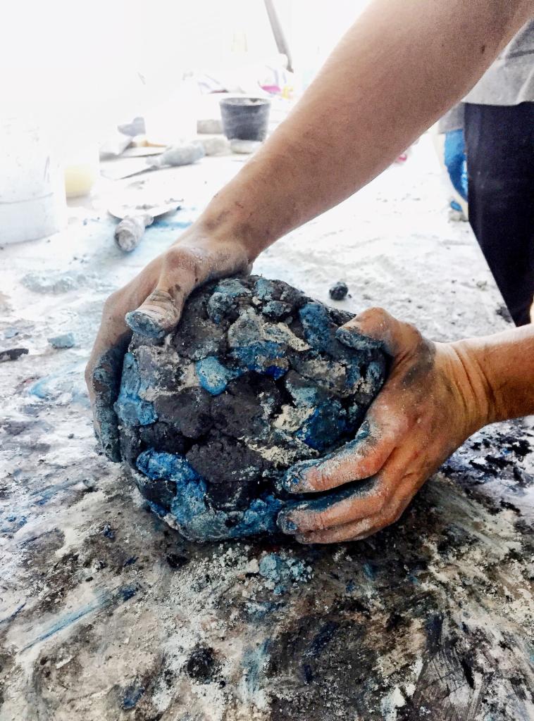Dłonie ugniatają kulę z szarej i niebieskiej gliny na pobrudzonym stole, w tle widoczne narzędzia i pojemniki.