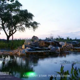 Kaskada wodna z kamieni w ogrodowym stawie oświetlona punktowymi lampami, widoczne lilie wodne i zielona roślinność, w tle drzewo i zachodzące słońce.