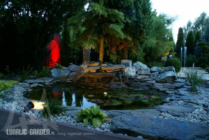 Oświetlony oczko wodne z kaskadą wodną i liliami wodnymi, otoczone kamieniami i roślinnością, z dekoracyjnym czerwonym elementem w tle i fontanną w oddali.