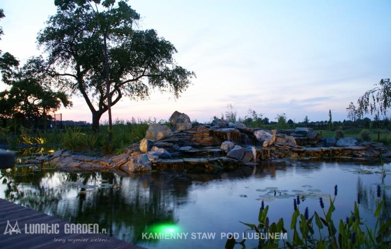 Kaskada wodna z kamieni w ogrodowym stawie oświetlona punktowymi lampami, widoczne lilie wodne i zielona roślinność, w tle drzewo i zachodzące słońce.