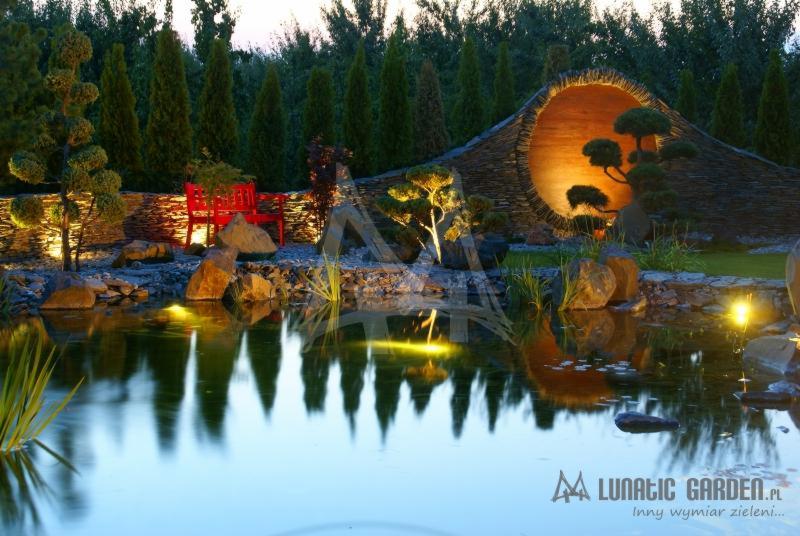 Oświetlony ogród z oczkiem wodnym, ozdobną ścianą z otworem i formowanymi drzewkami, odbicia w wodzie, czerwona ławka w tle.