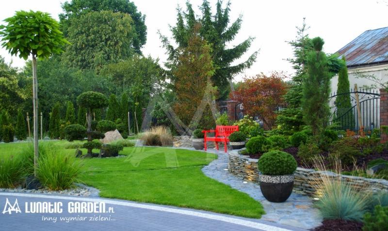Urzekający ogród z równo przystrzyżonym trawnikiem, ścieżką z kamienia prowadzącą do czerwonej ławki na tle muru z kamienia, ozdobne krzewy w donicach i różnorodne drzewa.