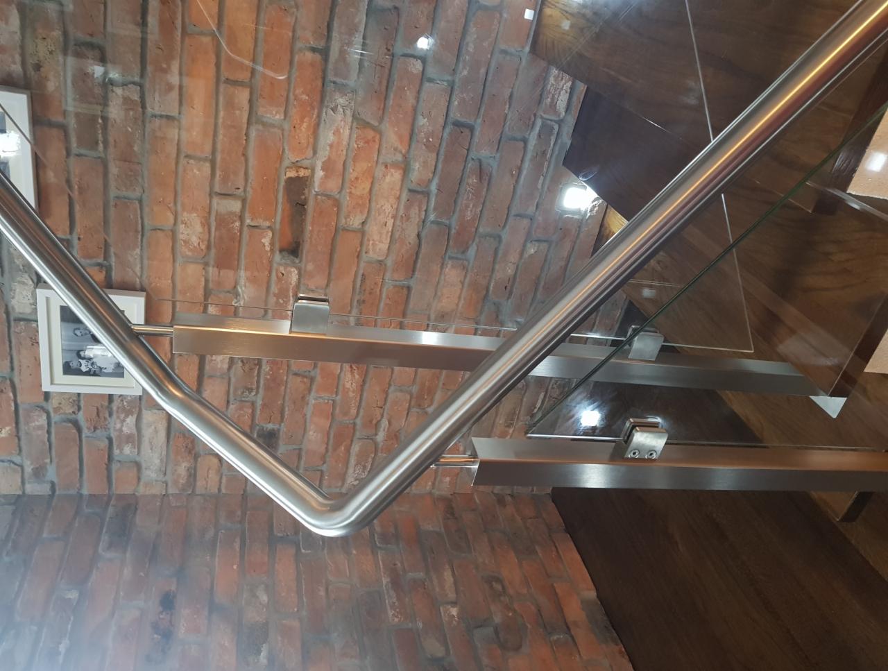 Szklana balustrada schodowa z metalowymi mocowaniami, widok z dołu, ceglana ściana w tle