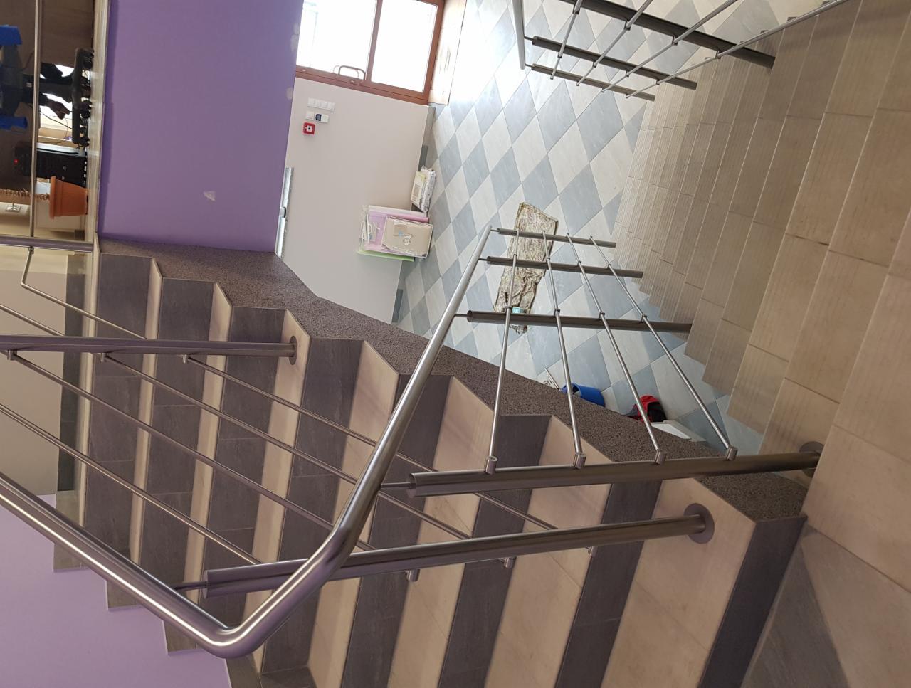 Schody wewnętrzne z metalową poręczą, widok z góry, szaro-fioletowe ściany i szachownica na podłodze przy oknie.