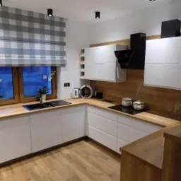 Nowoczesna kuchnia z białymi meblami na wymiar, drewnianym blatem i oknem z roletą w kratę, widok z perspektywy podłogi.