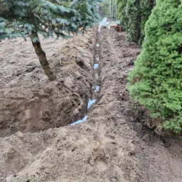 Wykopany rów w ziemi z widocznymi białymi rurami drenażowymi, obok rosną drzewa iglaste.