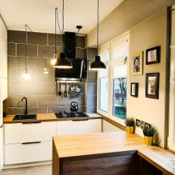 Nowoczesna kuchnia z szarymi płytkami na ścianie, białymi szafkami z czarnymi uchwytami, drewnianym blatem, czarnym zlewem i trzema wiszącymi lampami z czarnymi kloszami. Na ścianie obok okna wiszą...