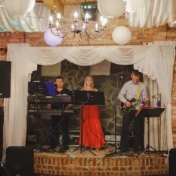 Trzyosobowy zespół na scenie z cegły, oświetlonej girlandami i lampionami, z wokalistką w czerwonej sukni, klawiszowcem i gitarzystą.