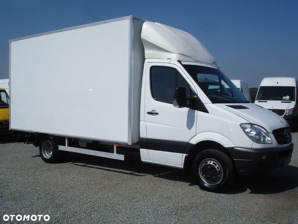 Biały samochód dostawczy Mercedes-Benz z zabudową kontenerową, zaparkowany w Płocku na żwirowym placu, widoczny z boku na tle innych pojazdów i błękitnego nieba.