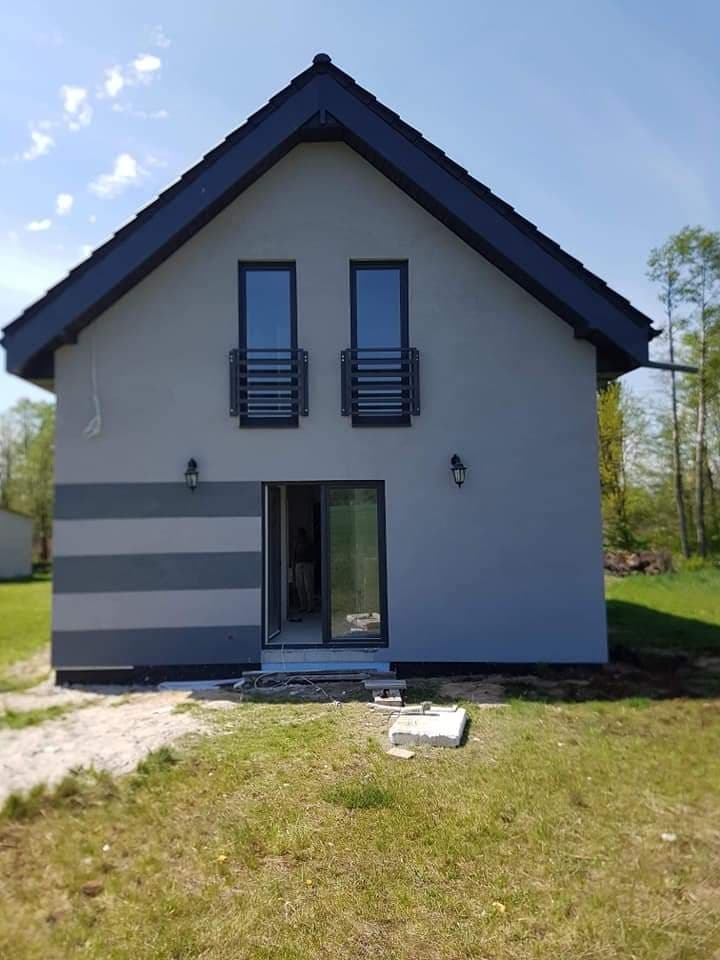 Elewacja domu jednorodzinnego w odcieniach szarości, z ciemnymi poziomymi pasami i czarną stolarką okienną, widok z zewnątrz na trawnik i niedokończone wejście.