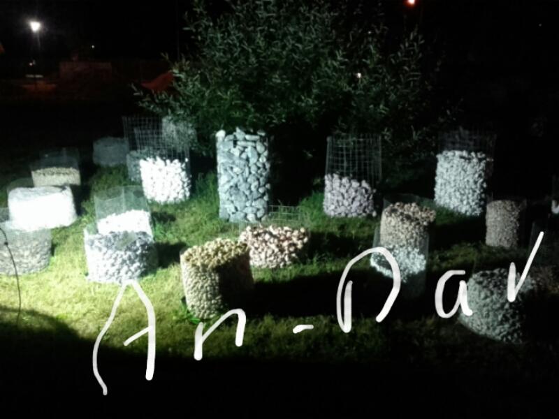 Nocne ujęcie dekoracyjnych gabionów wypełnionych różnymi rodzajami kamieni na trawniku, oświetlonych punktowo, z widocznym krzewem w tle.