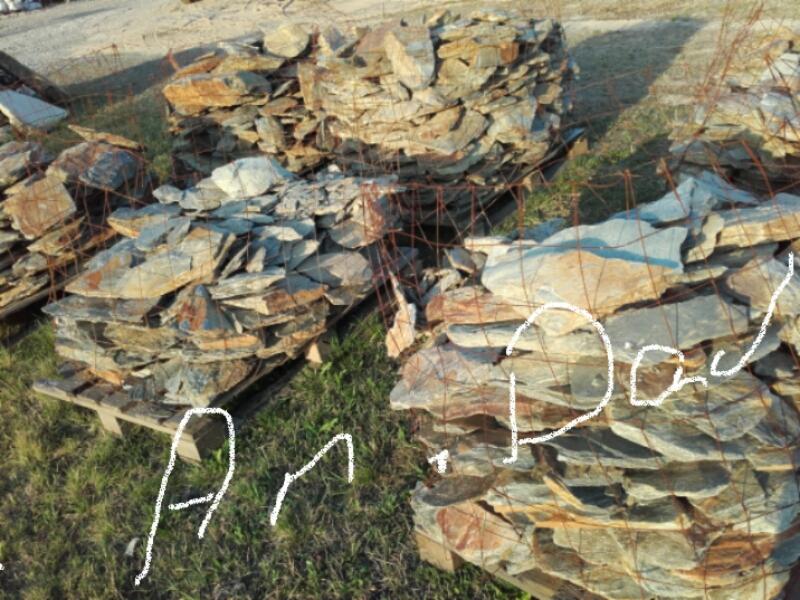 Palety z łupkiem kamiennym w metalowej siatce, widoczne trawa i piasek w tle.