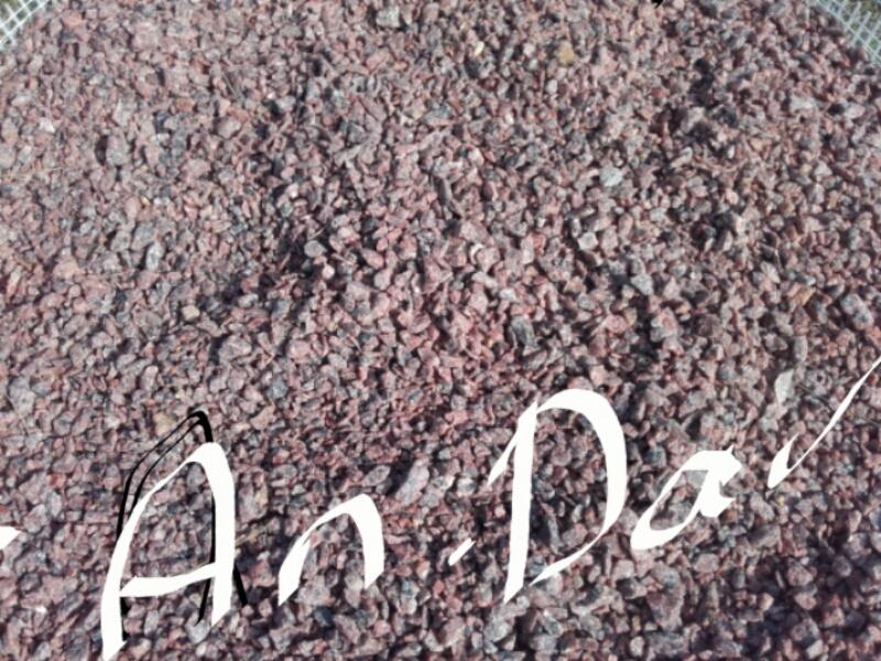 Czerwony grys granitowy w Jeleniej Górze, drobne kamyczki o nieregularnych kształtach, oświetlone naturalnym światłem, widoczny fragment siatki w rogu, tekst nałożony na zdjęcie.
