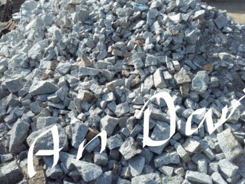 Góra szarego, drobnego kruszywa granitowego o nieregularnych kształtach, z widocznymi brązowymi przebarwieniami na niektórych kamieniach.