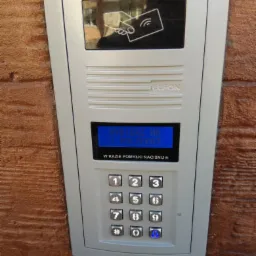 Panel wejściowy z klawiaturą numeryczną i czytnikiem kart zbliżeniowych, zamontowany na elewacji budynku o fakturze drewna.