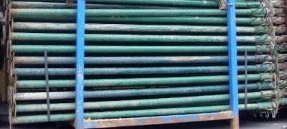 Stos zielonych, metalowych podpór budowlanych na niebieskim palecie, zabezpieczonych czarnymi pasami.