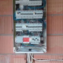 Otwarta skrzynka rozdzielcza z bezpiecznikami i przewodami na ścianie z czerwonej cegły, widoczne zabezpieczenia ETI i Eaton.