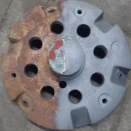 Okrągły metalowy element z otworami, w połowie pokryty rdzą, a w połowie szarą farbą, leżący na betonowej powierzchni.