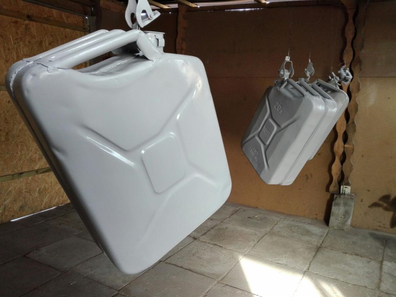 Dwie metalowe kanistry po obróbce, zawieszone na hakach w warsztacie. Jedna kanistra jest widoczna z bliska, ukazując gładką, szarą powłokę. Tło stanowią ściany z płyty OSB i betonowa podłoga.