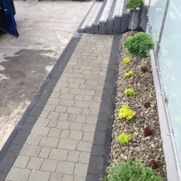 Ułożona ścieżka z kostki brukowej w odcieniach szarości, obramowana ciemniejszą kostką, prowadząca do schodów z tego samego materiału, z rabatą wysypaną korą i obsadzoną karłowatymi iglakami wzdłuż...