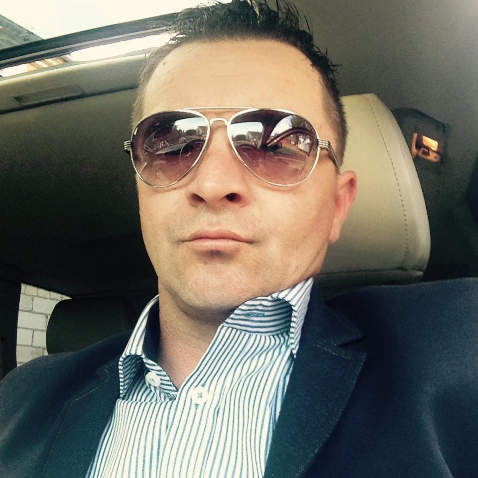 Portret mężczyzny w okularach przeciwsłonecznych, w samochodzie z widocznym fragmentem ceglanej ściany w tle.