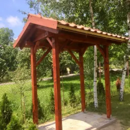 Drewniana konstrukcja z brązowym dachem krytym dachówką, wsparta na czterech słupach, umieszczona na betonowej podstawie, otoczona zielenią i drzewami.