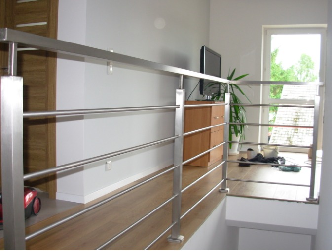 Nowoczesna, metalowa barierka schodowa z poziomymi prętami, widok z dołu, drewniana podłoga i drzwi w tle.