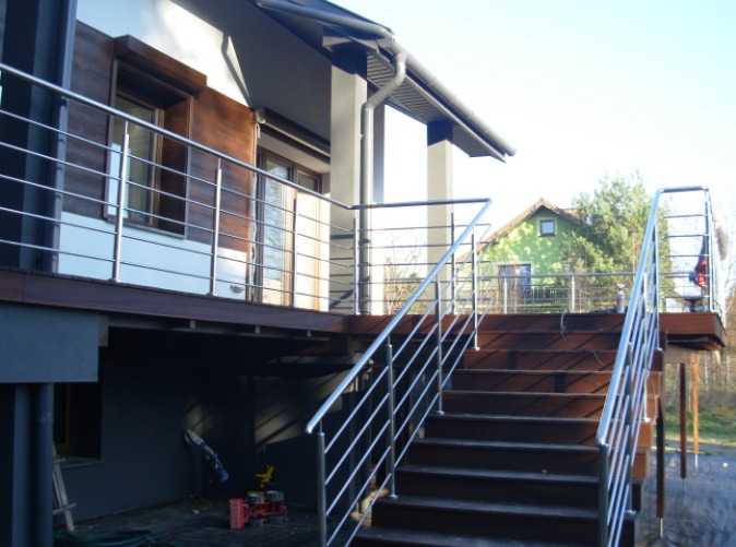 Zewnętrzne schody z drewnianymi stopniami i metalową balustradą z poziomych prętów, prowadzące na taras z podobną konstrukcją, w tle dom z drewnianą elewacją.