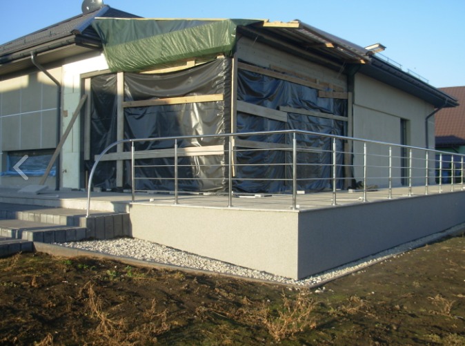 Nowoczesna, metalowa barierka balkonowa z rur na tle domu w trakcie budowy, zabezpieczonego czarną folią, widoczne schody wejściowe.