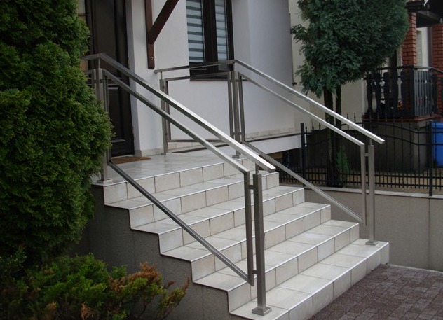 Zewnętrzne schody wyłożone jasnymi płytkami z zamontowanymi nowoczesnymi balustradami ze stali nierdzewnej o kwadratowych słupkach, prowadzące do drzwi wejściowych domu.