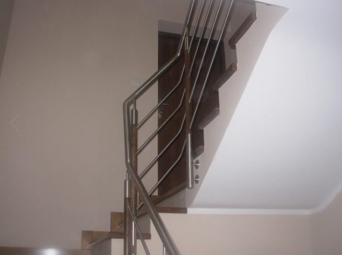 Klatka schodowa z drewnianymi stopniami i metalową, nowoczesną konstrukcją zabezpieczającą, zamontowaną przy ścianie w jasnym wnętrzu.