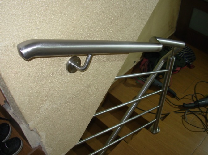 Szczegółowe ujęcie montażu poręczy ze stali nierdzewnej przy schodach, widoczne mocowanie do ściany i podłogi, w tle narzędzia i ubrania.