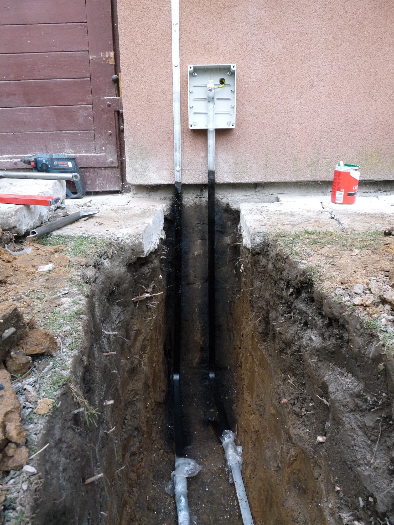 Wykop w ziemi z ułożonymi rurami i puszką elektryczną na ścianie budynku, widoczne narzędzia i materiały budowlane.