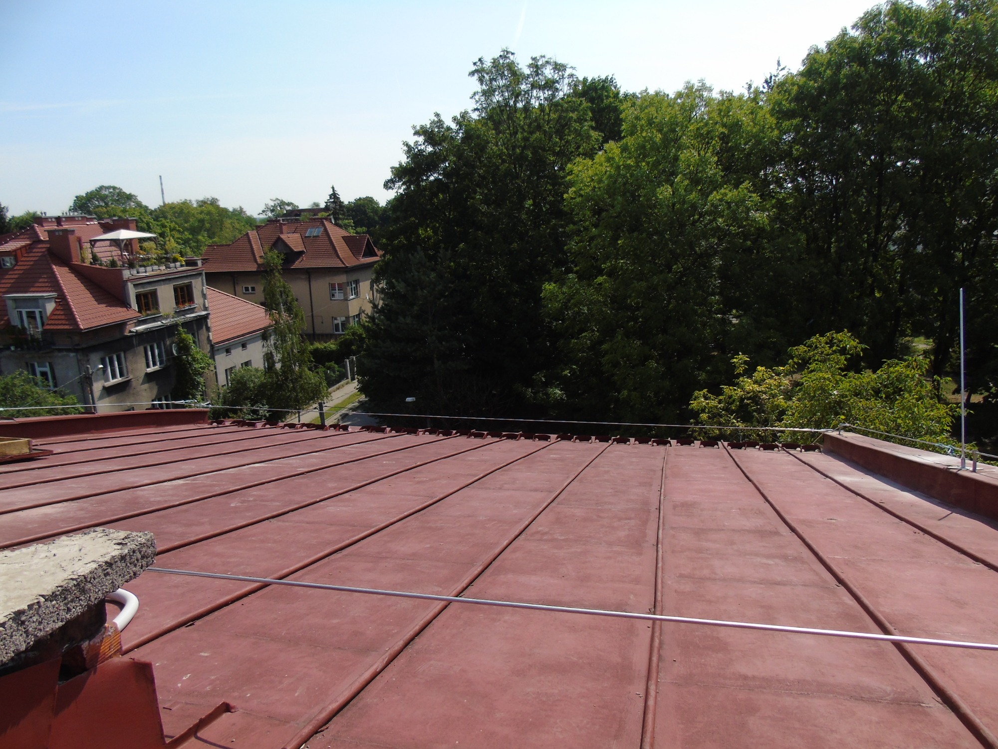 Czerwony blaszany dach z widocznymi elementami instalacji odgromowej, w tle zabudowa miejska i zieleń drzew.
