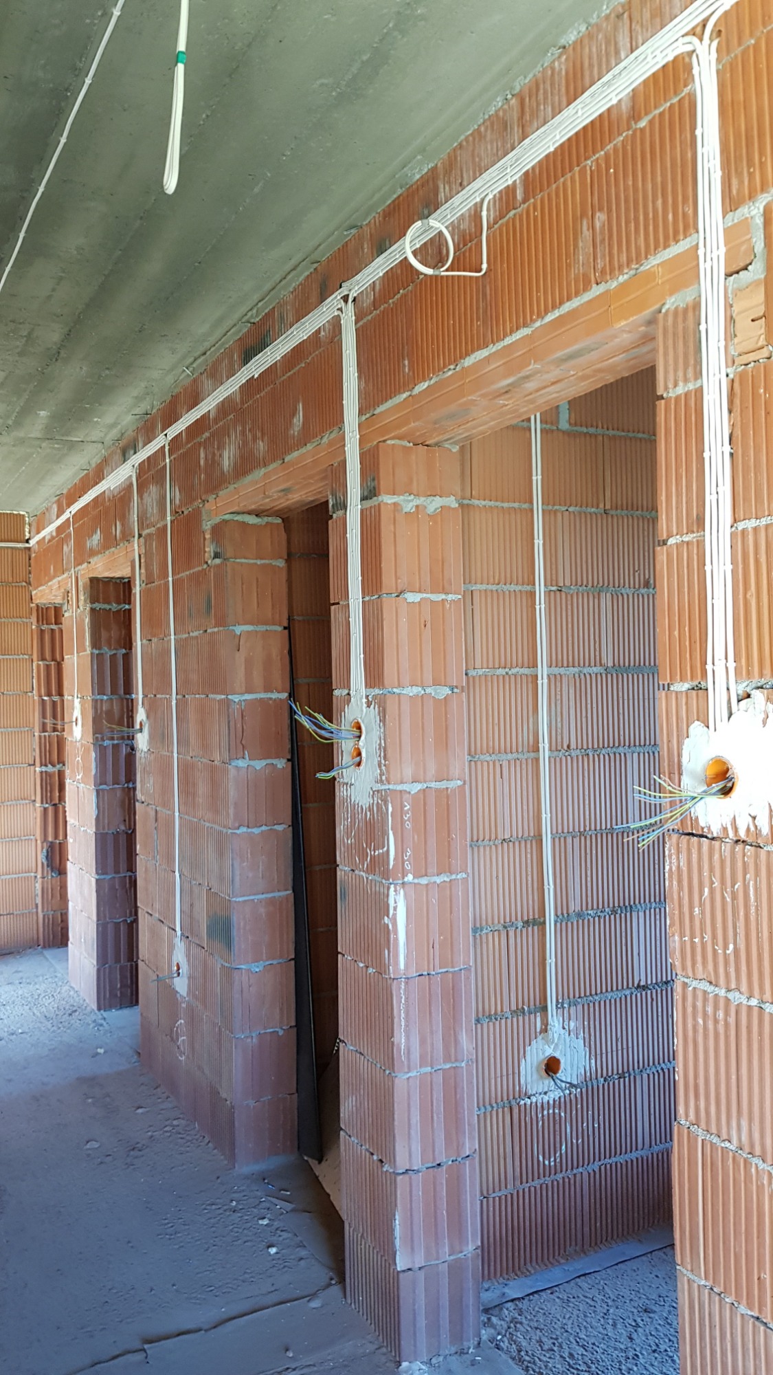 Wewnętrzne instalacje elektryczne w surowym budynku z czerwonej cegły, widoczne kable w białych peszlach i puszki instalacyjne z wystającymi przewodami.