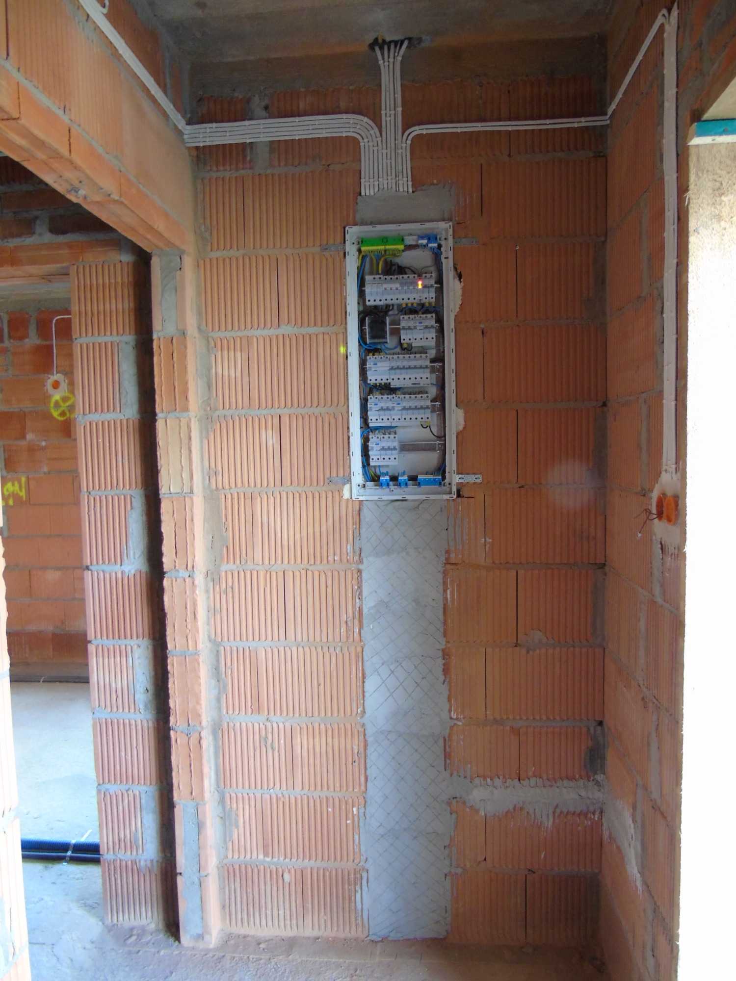 Rozdzielnica elektryczna zamontowana na ścianie z czerwonej cegły, przewody prowadzone w korytkach, widoczne ślady po szpachlowaniu ściany pod tynk.