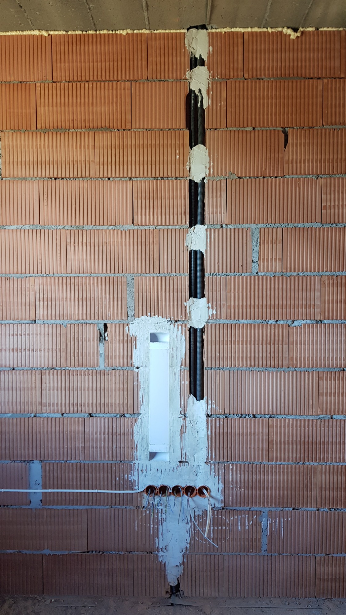 Pionowy peszel elektryczny zamocowany w ścianie z cegły dziurawki, zabezpieczony gipsem, widoczne puszki instalacyjne z przewodami.