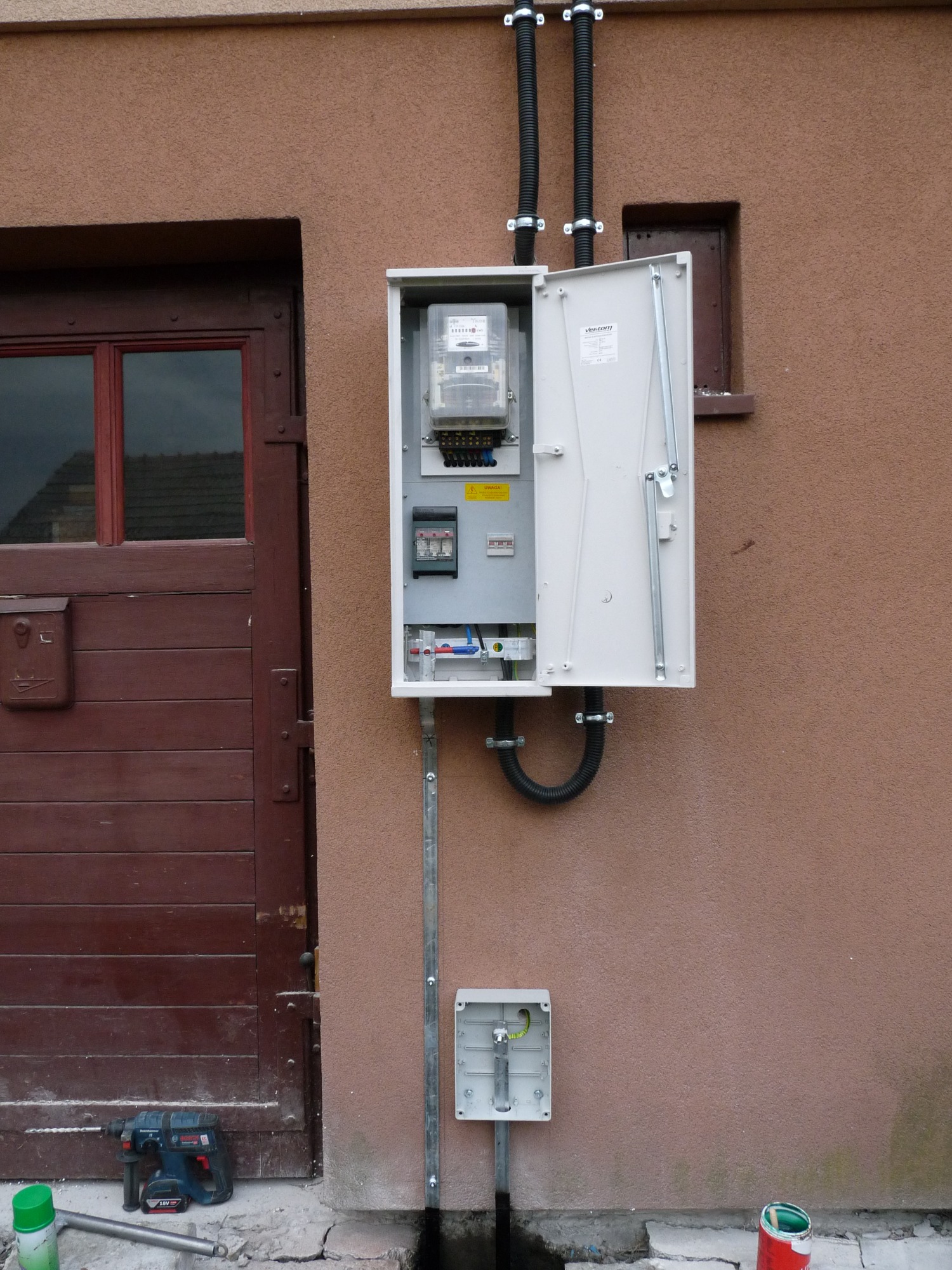 Otwarta skrzynka z licznikiem elektrycznym zamontowana na ścianie budynku, widoczne przewody w peszlach, dodatkowa puszka instalacyjna poniżej, w tle brązowe drzwi i ściana, na ziemi wiertarka...