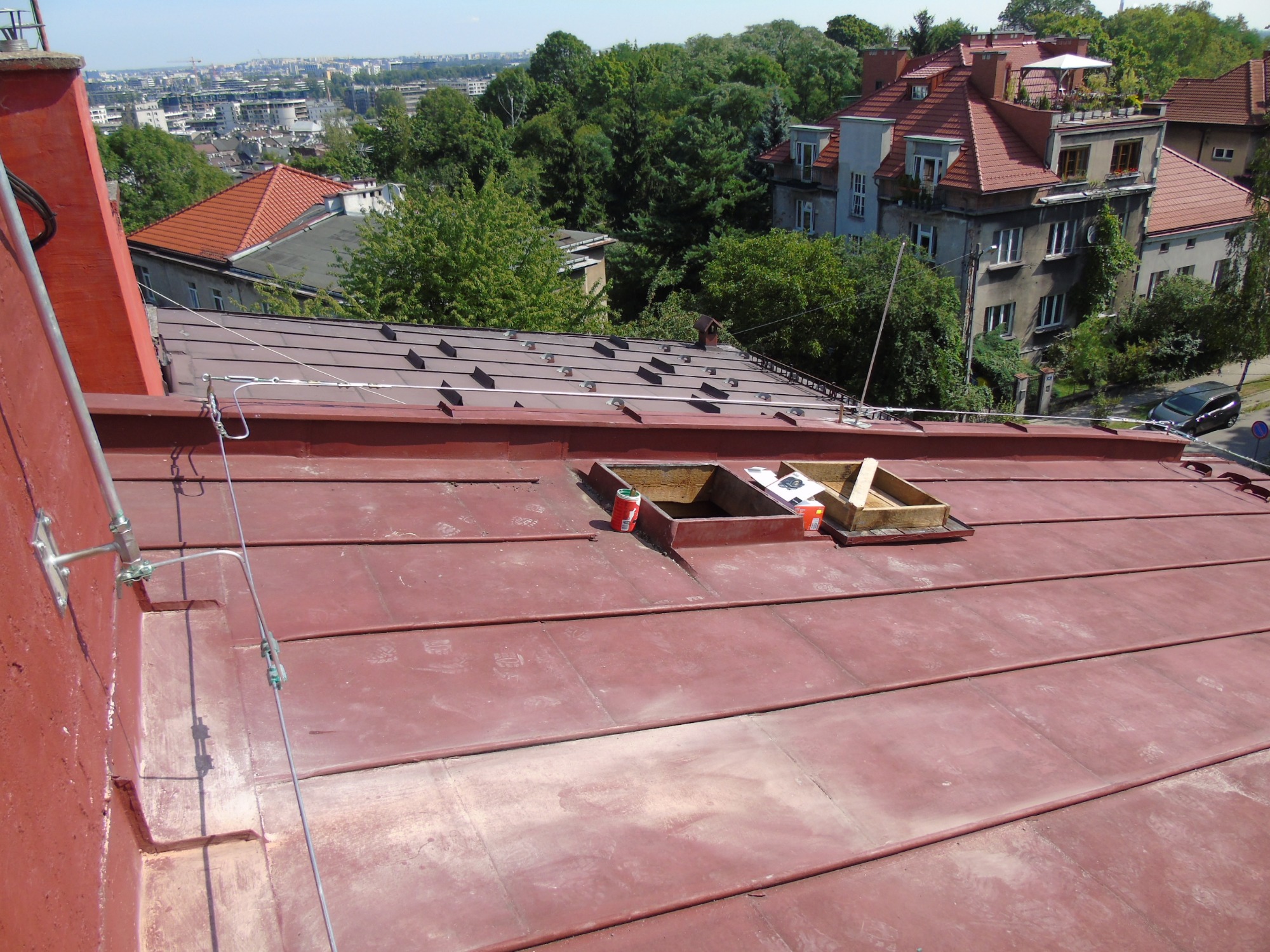 Czerwony blaszany dach z widocznymi elementami instalacji odgromowej, otworami wentylacyjnymi i puszkami farby, w tle zabudowa miejska i zieleń.