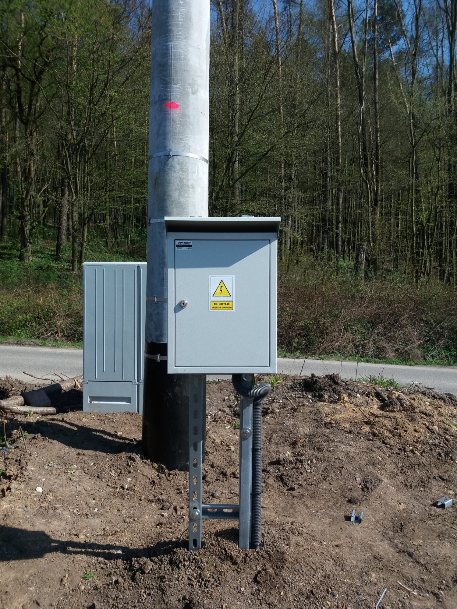 Szara skrzynka elektryczna z ostrzeżeniem o napięciu, zamontowana na słupie oświetleniowym z metalowymi wspornikami, na tle lasu i drogi.