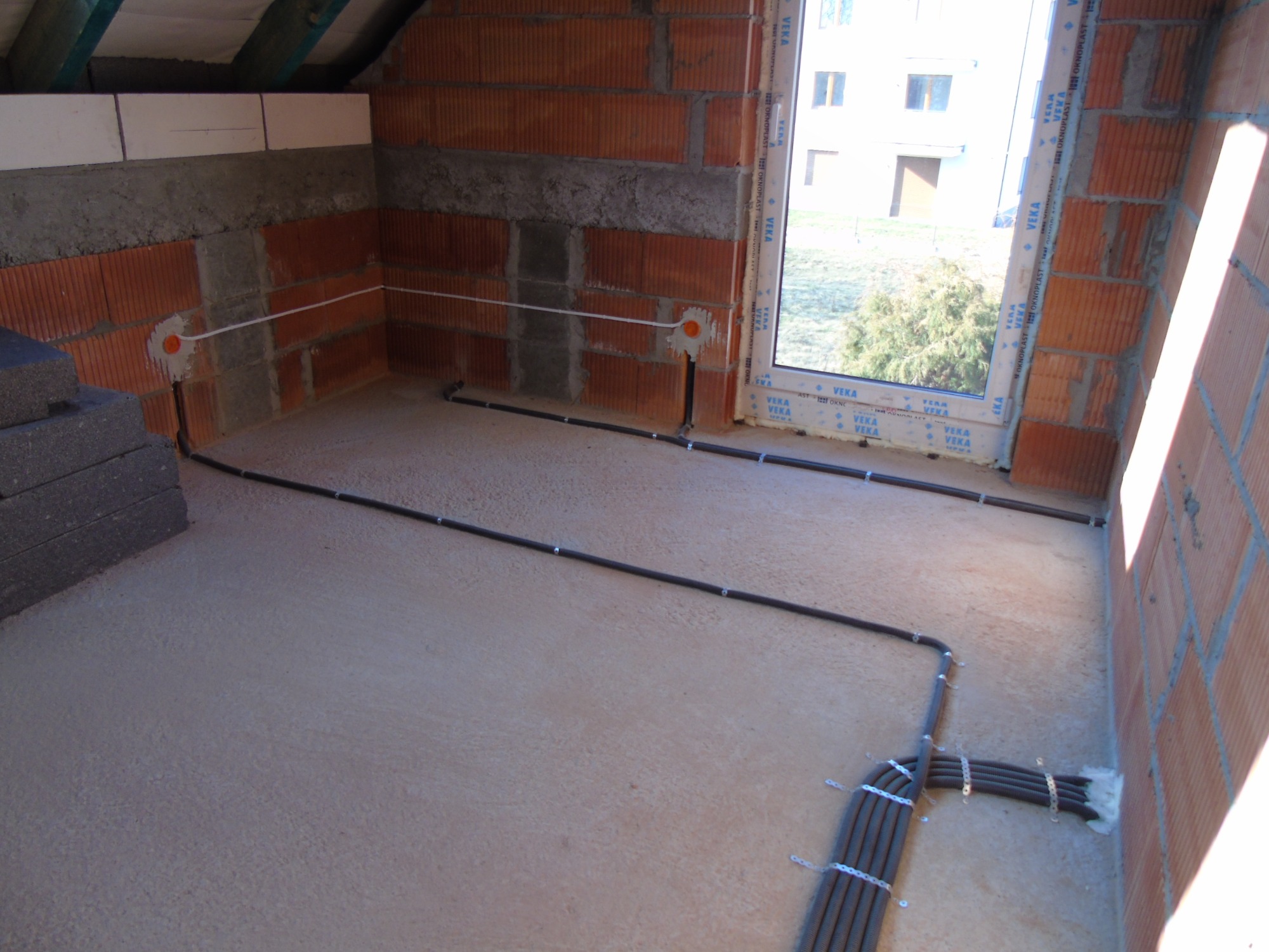 Wewnętrzna instalacja elektryczna w trakcie montażu w surowym budynku: kable prowadzone po podłodze i ścianach, mocowane klamrami, widoczne puszki instalacyjne w ścianie z cegły dziurawki, okno...