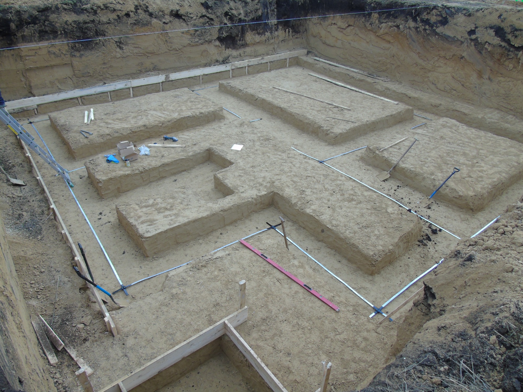 Wykop fundamentów z ułożonymi bednarkami i szalunkami, widoczne narzędzia budowlane i poziomica w wykopie.