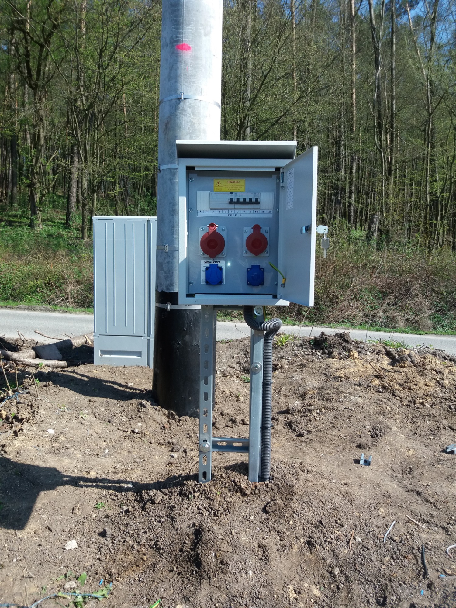 Słup oświetleniowy z otwartą skrzynką elektryczną zawierającą gniazda siłowe i bezpieczniki, zamontowany na metalowej konstrukcji w ziemi, na tle lasu.