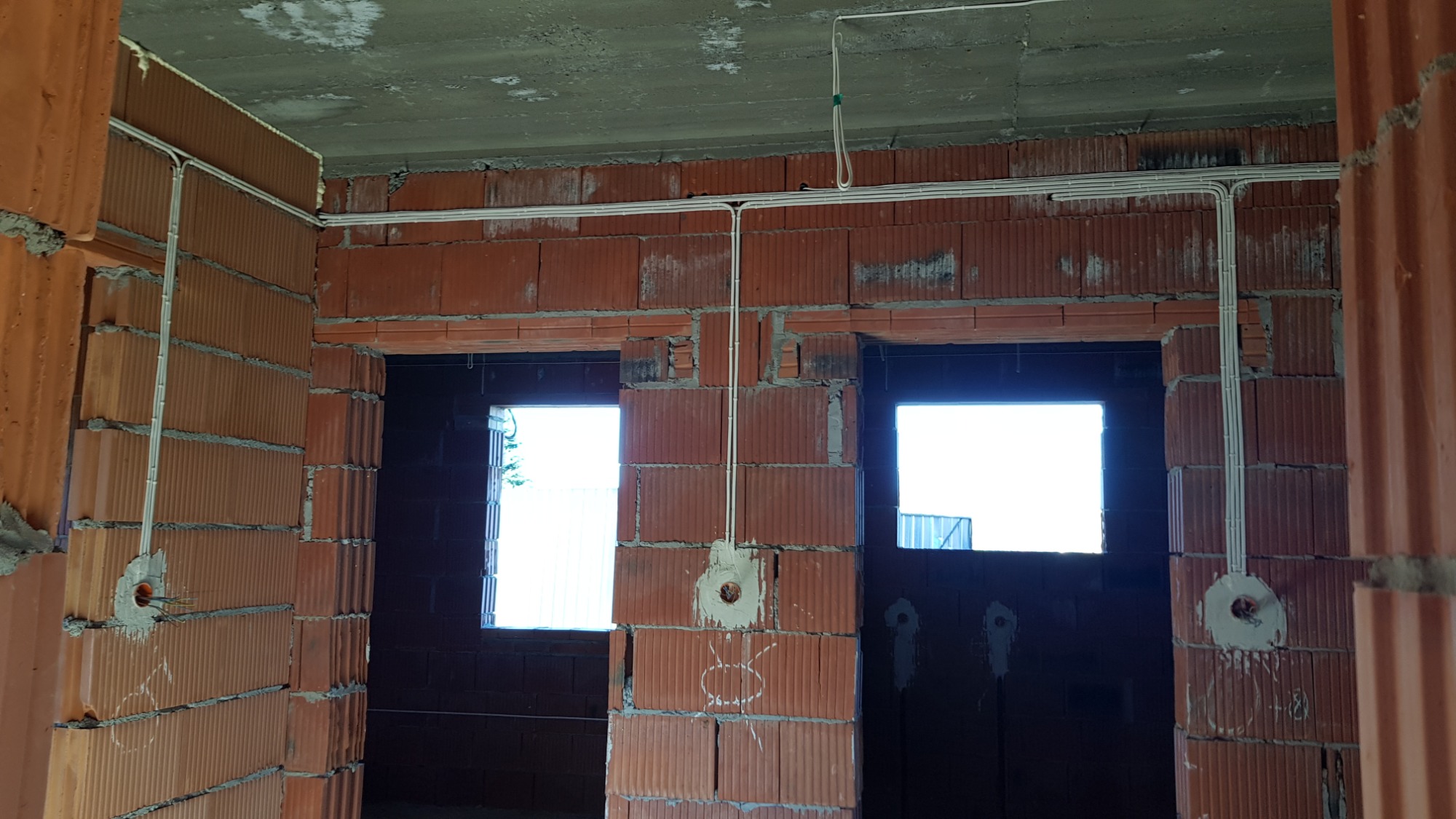 Surowe wnętrze w trakcie budowy z widocznymi przewodami elektrycznymi poprowadzonymi po ścianach z czerwonej cegły dziurawki oraz przygotowanymi otworami pod gniazdka.