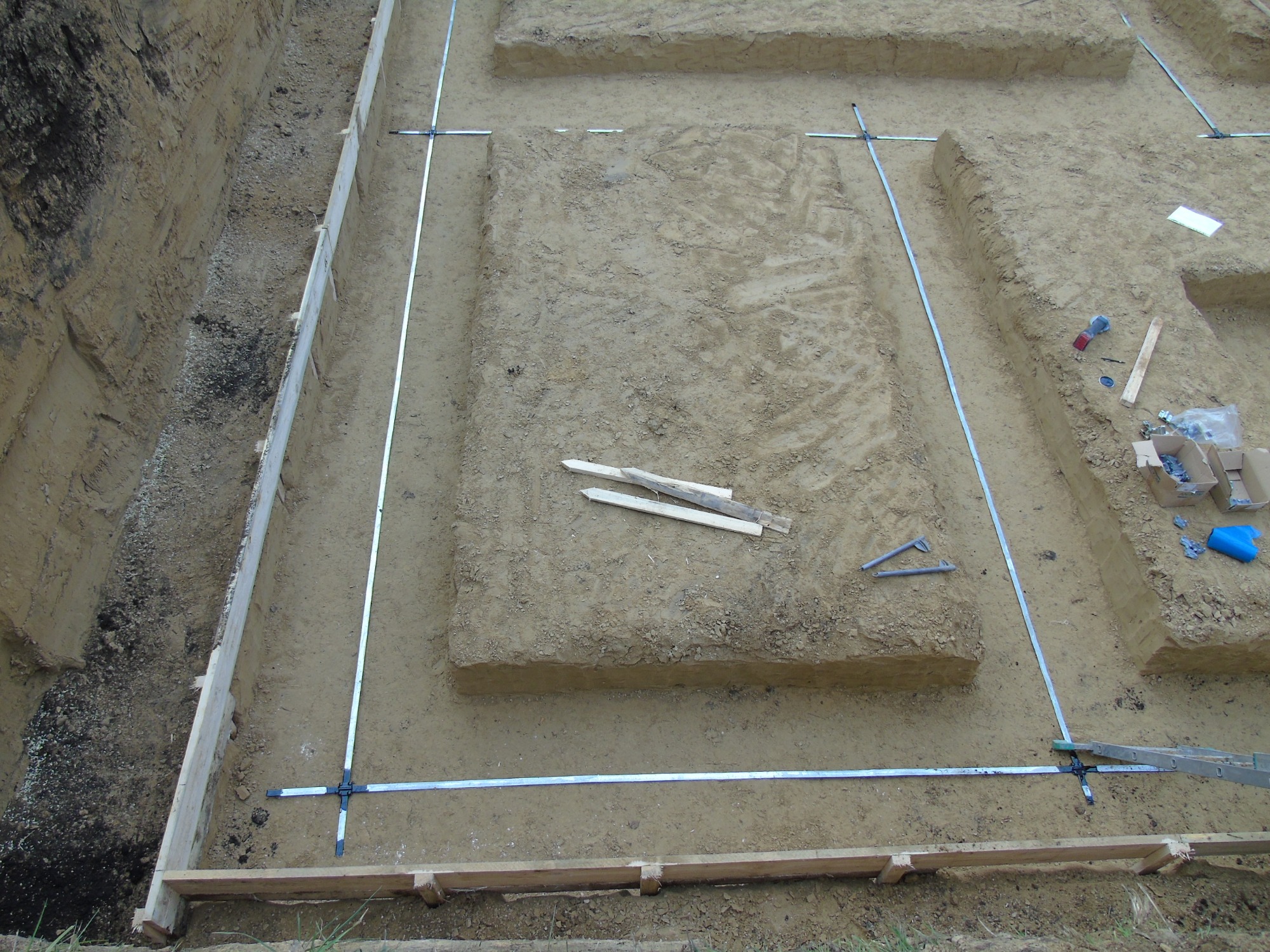 Wykonane wykopy pod fundamenty domu, widoczne szalunki i metalowe profile wyznaczające obrys, ziemia piaszczysta, narzędzia i materiały budowlane w tle.