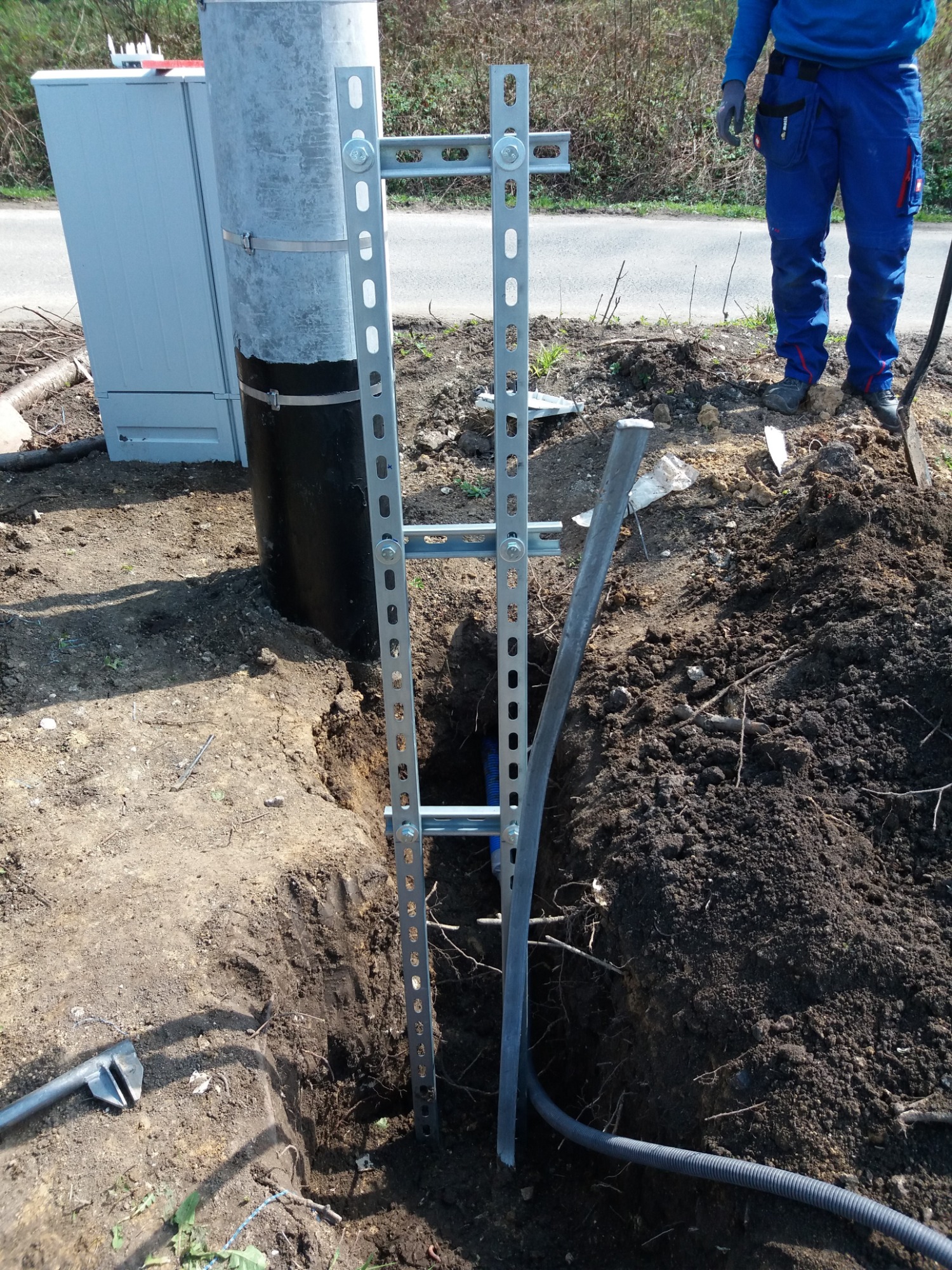 Słup energetyczny z zamontowaną metalową konstrukcją wsporczą w wykopie, widoczny kabel w karbowanej osłonie i częściowo widoczna szafka rozdzielcza obok.